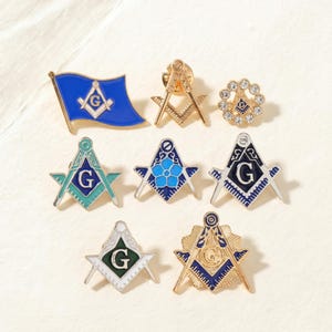 May include: A collection of Masonic pins in various designs. The pins feature symbols like the square and compass, a flag, and the letter "G". Colors include gold, blue, green, and white. The pins are likely made of metal and have a pin back.