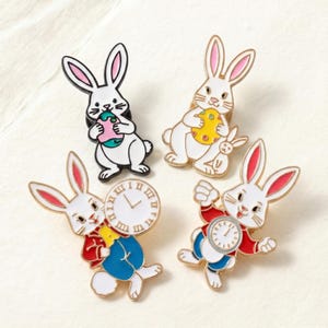 May include: Four enamel pins featuring white rabbits with pink-lined ears. Two rabbits hold Easter eggs, one with a pink and teal egg, the other with a yellow egg. The other two rabbits wear red jackets and blue pants, holding white clocks.