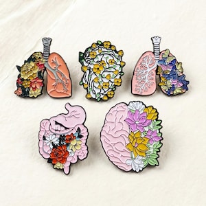 Könnte beinhalten: Eine Sammlung von fünf Emaille-Anstecknadeln mit anatomischen Designs. Die Anstecknadeln zeigen Lungen, ein Gehirn und einen Darm, jeweils mit bunten Blumen- und Schmetterlingsakzenten verziert. Die Anstecknadeln sind vor einem neutralen Hintergrund platziert.