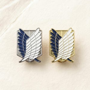 Peut inclure: Deux épingles métalliques avec un motif d'ailes stylisées. L'une est argentée avec de l'émail bleu et blanc, l'autre dorée avec de l'émail bleu et blanc. Ces épingles sont pour vêtements ou accessoires.