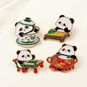 May include: Four enamel panda pins. One panda is in a teacup, another is playing Go, a third is playing mahjong, and the last is in a hot pot. Each pin has gold-colored trim and is set against a white background.