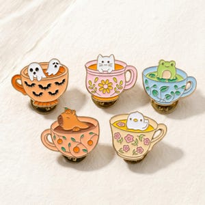 May include: Set of five enamel pins featuring cute animals and objects in teacups. Designs include a ghost, cat, frog, capybara, and duck. Each pin has a unique color scheme, including orange, pink, blue, and yellow, with gold-colored metal accents.