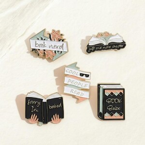 May include: A selection of enamel pins with book-related designs. The pins feature phrases such as "book nerd," "just one more chapter," and "cool people read." The pins are in various colours, including white, black, pink, and mint green.
