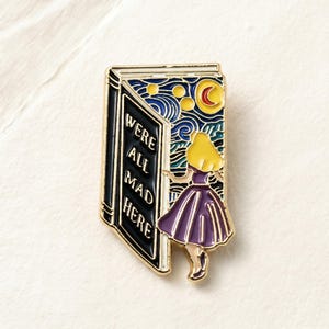 May include: Enamel pin featuring a scene from Alice in Wonderland. The pin depicts a book with the words "WE'RE ALL MAD HERE" and a character in a purple dress. The background is a starry night sky with a crescent moon.