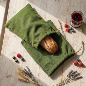 May include: A green linen bread bag with a drawstring closure, holding a loaf of bread. A jar of jam, berries, and sprigs of lavender and wheat are arranged on a rustic wooden surface, creating a charming still life scene.