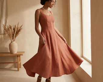 Puede incluir: Un vestido de lino color óxido con escote cuadrado y falda evasé. El vestido sin mangas se combina con sandalias de cuña blancas. La modelo está de pie cerca de una ventana.