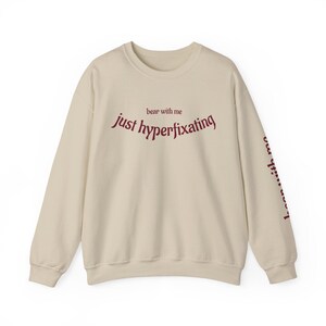 May include: A cream-coloured sweatshirt featuring the burgundy text "bear with me just hyperfixating" in a curved font across the chest. The same text is repeated on the left sleeve. The sweatshirt has a crew neck and long sleeves.