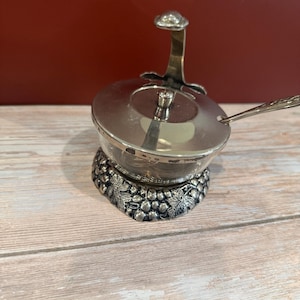 May include: A silver-colored serving dish with a lid and a small spoon. The dish sits on a decorative base with a leaf design. The handle on the lid has a decorative finial. The background is a wooden surface.