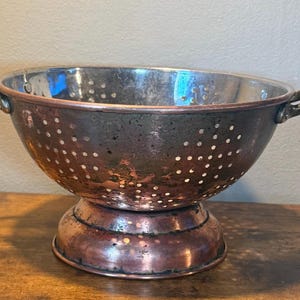 May include: A round copper colander featuring a metallic finish with visible patina, two side handles, and a pedestal base. The bowl is perforated with numerous small circular holes throughout the sides and base for draining liquids from food items.