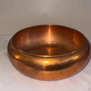 May include: A round, shallow copper bowl with a polished metallic finish. The bowl features a smooth, curved exterior wall and a wide, open interior basin. The surface displays subtle variations in color and light reflections across the metal.