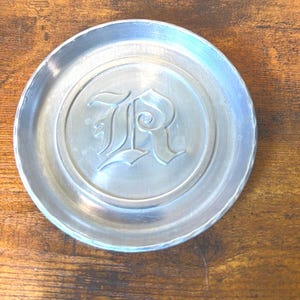May include: A circular silver-toned metal coaster featuring a raised, embossed letter R in an ornate gothic font centered within a smooth circular border. The coaster has a slightly hammered or textured edge and a polished metallic finish.
