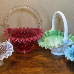 May include: A collection of four glass baskets featuring hobnail patterns and ruffled edges. The set includes a clear glass basket, a deep red glass basket, a white and light green ombre glass basket, and a bright blue glass basket.