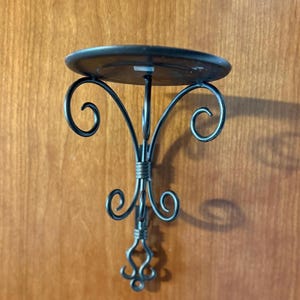 May include: A black metal wall-mounted candle holder featuring a circular top platform supported by a decorative frame with curved scrollwork details and a central vertical bar that terminates in a small, intricate ornamental base.