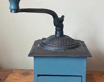 May include: A blue wooden coffee grinder featuring a black cast iron top with intricate embossed floral patterns, a black metal crank handle, and a matching blue wooden knob. The base includes a small pull-out drawer with a round blue wooden knob.