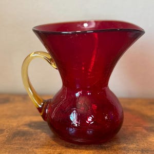 May include: A deep red glass pitcher featuring a distinct crackle texture throughout the body. The vessel has a wide, flared rim and a curved, translucent amber-colored glass handle attached to the side, creating a contrast in color and form.