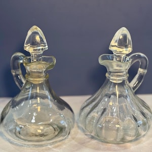 May include: Two clear glass cruet bottles with handles and cone-shaped stoppers. The bottles have a bulbous shape with vertical ridges. The background is a dark blue.