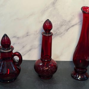 May include: Three ruby red glass bottles of varying shapes. The bottle on the left is a small jug with a handle and a stopper. The middle bottle is tall and cylindrical with a decorative base. The bottle on the right is a tall vase.