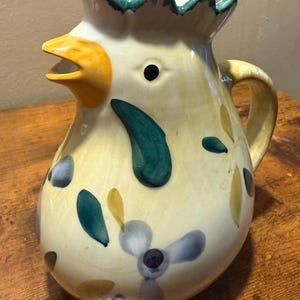 May include: A ceramic pitcher shaped like a chicken with a yellow body, a bright orange beak, and a green comb. The surface is decorated with hand-painted teal and blue leaf patterns and floral motifs. It features a curved handle and a wide spout.