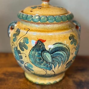 May include: A yellow ceramic jar with a lid, featuring a hand-painted teal rooster with a red comb. The jar has two side handles, a textured green rim, and green leaf motifs painted around the body. The glaze has a glossy, mottled finish.