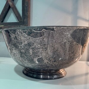 May include: A large, silver-toned bowl with a textured surface and a pedestal base. The bowl has a wide opening and a rounded shape, suitable for serving or display. The bowl's exterior has a marbled appearance.