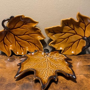 May include: Three ceramic dishes shaped like autumn leaves in a glossy mustard yellow glaze. Each dish features dark brown, raised vein detailing and a dark brown stem accent. The dishes have a smooth, reflective finish and irregular edges.