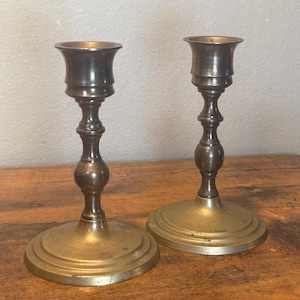 May include: Two matching metallic candle holders with a polished finish. Each piece features a wide circular base, a slender stem with a central rounded bulbous detail, and a flared top cup designed to securely hold a standard taper candle.