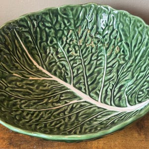 May include: A large, round ceramic bowl shaped like a cabbage leaf. The bowl features a glossy, deep green finish with intricate, raised textures mimicking the veins and crinkled surface of a cabbage leaf, accented by a prominent white central vein.