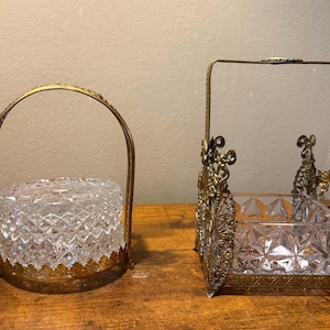 May include: A round clear cut glass bowl with a gold-toned metal handle and base, alongside a square clear cut glass bowl featuring an ornate gold-toned metal frame with a tall handle and decorative filigree corner accents on a wooden surface.