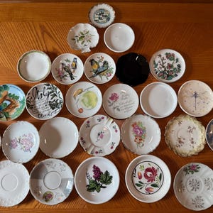 May include: A collection of twenty-four small ceramic saucers and dishes arranged in a triangular formation. The pieces feature various designs including floral patterns, bird illustrations, landscape scenes, fruit motifs, and solid white surfaces.