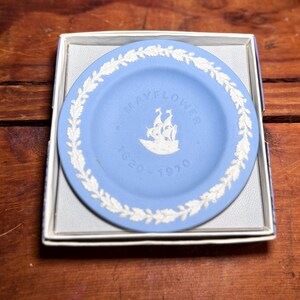 May include: A light blue circular commemorative plate featuring a white relief of the Mayflower ship, the text MAYFLOWER, and the years 1620-1970. The plate is bordered by a white leaf wreath pattern and is presented inside a square white box.