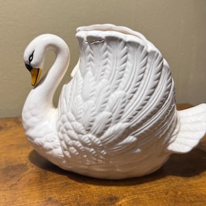 May include: A white ceramic swan-shaped planter or decorative vessel featuring molded feather details on the body and wings. The swan has a curved neck, a yellow and black beak, and black eye markings. The top of the swan's back is open.