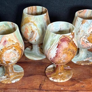 May include: A set of four goblets crafted from polished banded stone, featuring a rounded bowl, a short stem, and a circular base. Each goblet displays unique natural patterns in shades of sage green, cream, amber, and deep reddish-brown.