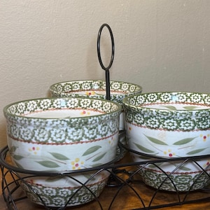 May include: A set of three ceramic bowls arranged in a black metal wire caddy with a central carrying handle. Each bowl features a white base with a green floral pattern, small red and yellow accents, and a decorative green border along the rim.