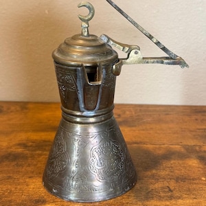 May include: A metallic coffee pot with a dark, tarnished finish featuring intricate engraved patterns on the body. The lid is topped with a crescent moon finial, and a long, angled metal handle extends from the side, attached by a hinged mechanism.