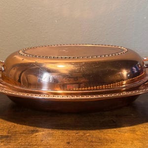 May include: An oval copper serving dish with a matching lid, featuring a polished metallic finish and decorative beaded edges along the rim and lid. The piece includes two small, curved handles on opposite sides for lifting and transport.
