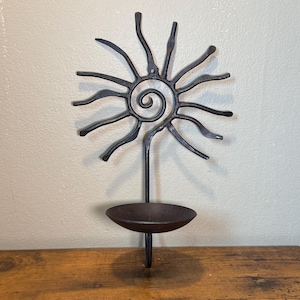 May include: A dark metal wall-mounted candle holder featuring a sun-shaped design with a central spiral and radiating curved rays. A single circular bowl-shaped platform extends from the bottom of the sun motif to hold a candle securely.