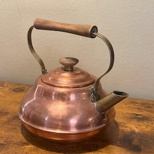 May include: A copper tea kettle with a wide, rounded base and a short, angled spout. The kettle features a brass handle with a dark brown wooden grip and a matching wooden knob on the removable lid. The metal surface has a polished finish.