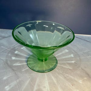 May include: A green glass sundae dish with a flared bowl and a pedestal base. The bowl has vertical ridges, and the glass has a slight green tint. The dish is sitting on a light-colored surface.