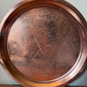 May include: A round copper tray featuring an intricate engraved floral pattern across its surface. The metallic finish has a warm reddish-brown tone with visible light reflections and subtle surface scratches throughout the circular metal plate.
