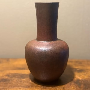 May include: A tall, bulbous vase crafted from dark, reddish-brown metal. The surface features a distinct hammered texture throughout, creating a matte finish. The vase has a narrow, cylindrical neck that widens into a rounded, spherical body.