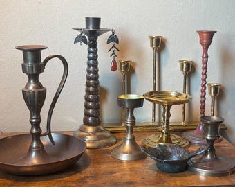 May include: A collection of various metal candle holders in different shapes and sizes, including a large copper-toned chamberstick with a handle, a tall stacked-sphere holder, several brass pillar stands, and a small dark textured bowl.