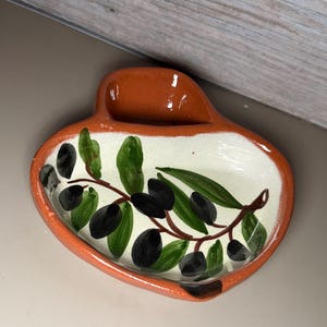 May include: Ceramic olive dish with a terracotta rim and handle. The interior is white with a painted olive branch design featuring green leaves and black olives. A decorative piece for serving olives.