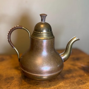 May include: A copper teapot with a rounded body, a brass lid featuring a small wooden knob, and a curved brass spout. The handle is made of brass and wrapped in a dark brown textured material. The metal surfaces show a natural patina finish.