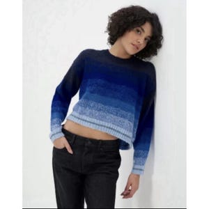 Puede incluir: Un suéter azul degradado con cuello redondo y mangas largas. El suéter pasa del azul oscuro a tonos más claros de azul. El dobladillo inferior y los puños tienen una raya azul claro y verde. Una prenda de punto elegante.