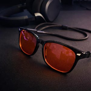 May include: Black-framed glasses with orange-tinted lenses, resting on a dark surface. The glasses have a cord attached. A pair of headphones and a keyboard are visible in the background. Gaming accessories.