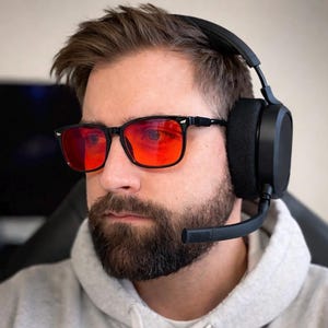 May include: A person wearing black-framed glasses with red lenses and a black gaming headset with a microphone. The glasses have a rectangular shape. The headset has large earcups and a flexible microphone boom.