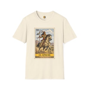 May include: A cream-colored short-sleeve t-shirt featuring a central graphic print of a lottery card design. The graphic depicts a person riding a spotted horse, holding a spear, with the number 38 and the text EL APACHE printed below.
