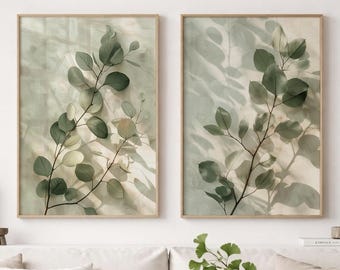 May include: Two rectangular wall art prints featuring green eucalyptus branches with soft shadows on a light background, displayed in thin wooden frames. The artwork depicts botanical foliage with layered leaves in varying shades of green.