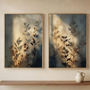 May include: Two rectangular wall art prints in light wood frames featuring a botanical motif with dark silhouettes of leafy branches against a soft, hazy background of golden light and deep blue shadows, displayed side by side on a wall.
