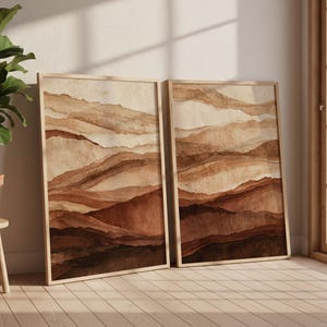 May include: Two rectangular framed wall art prints featuring abstract layered landscape designs in shades of beige, tan, and dark brown. The artwork displays horizontal, wavy, and textured bands that mimic natural terrain.